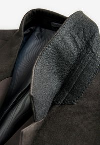 Colletto di un blazer nero con un morbido rivestimento grigio testurizzato. Presenta un tessuto interno nero liscio con un sottile motivo a righe.