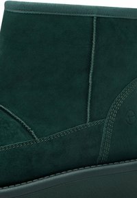 Stivaletto in suede verde con cuciture a contrasto, forma arrotondata e finitura testurizzata. Presenta una suola in gomma resistente e un dettaglio di branding discreto.