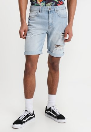 Shorts en denim bleu clair avec des ourlets retroussés, présentant des déchirures et des franges. Associés à une chemise à imprimé tropical et des baskets noires.