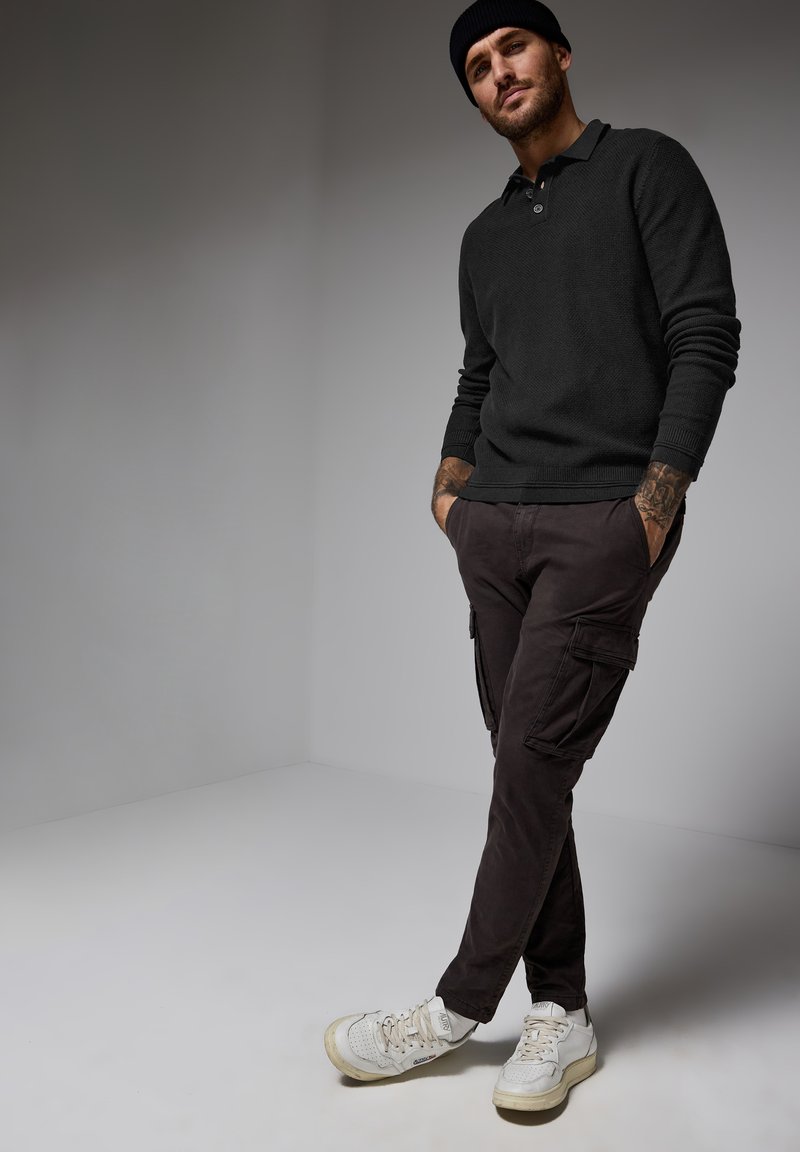 Svart stickad tröja med krage, mörka cargobyxor med sidofickor och vita sneakers. Modellen står med händerna i fickorna mot en grå bakgrund.