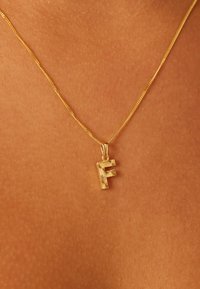 Gouden halsketting met een gestileerde hanger in de vorm van de letter "F" met gestructureerde details, hangend aan een fijne ketting tegen een huidskleurige achtergrond.