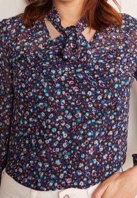 Blusa floreale di colore blu scuro con motivi multicolore vivaci. Presenta un design con nodo al collo e un tessuto trasparente con una sottile texture.