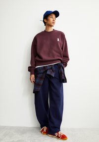 Bordeauxfarbener Pullover mit weißem Logo, marineblaue weite Hosen, karierte Hemd um die Taille gebunden, rote Sneakers mit beigen Schnürsenkeln, blauer Hut.