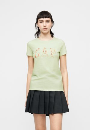 Giovane donna con capelli scuri indossa una T-shirt verde chiaro con il logo "GAP" floreale e una gonna scura plissettata su sfondo neutro.