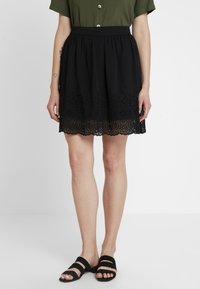 Svart kjol med broderad blommönster längs fållen, elastisk midja och en svävande silhuett, tillsammans med svarta sandaler.