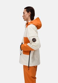 Chaqueta impermeable blanca con acentos naranjas, capucha ajustable, bolsillos laterales y un parche con un logo en la manga; combinada con pantalones naranjas.
