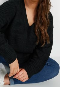 Kvinna med långt brunt hår sitter i kors med benen, iklädd en svart stickad tröja, blå jeans och vita sneakers, medan hon justerar sin manschett.