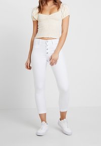 Białe, wysokiej talii jeansy o wąskim kroju i skróconej długości, z zapięciem na guziki. Połączone z żółtym, krótkim topem w paski.