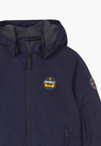 Ciemnoniebieska kurtka z kapturem, z miękkim szarym polarem wewnątrz, z naszywką z logo Ninjago na klatce piersiowej oraz okrągłą naszywką na rękawie.