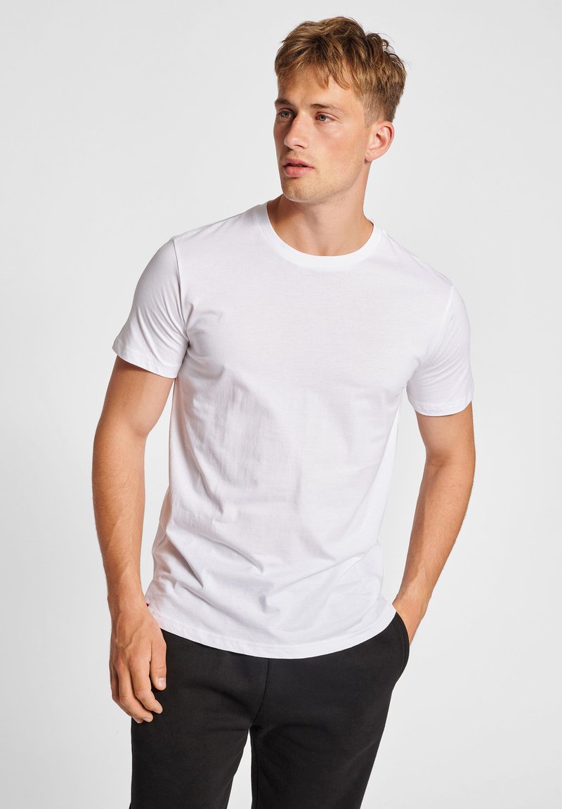 Junger Mann mit kurzen blonden Haaren, der ein einfaches weißes T-Shirt und schwarze Hose trägt, steht mit einer Hand in der Tasche und schaut zur Seite.