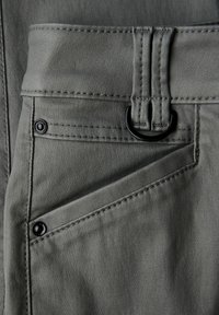 Gros plan d’une poche de pantalon en tissu gris avec des rivets noirs et un anneau en D noir fixé à une boucle de ceinture doublement cousue.