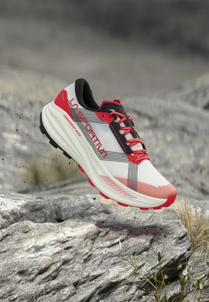 Λευκό και κόκκινο παπούτσι τρεξίματος La Sportiva κρέμεται πάνω από βραχώδες έδαφος με γρασίδι και μικρά λουλούδια στο βάθος.