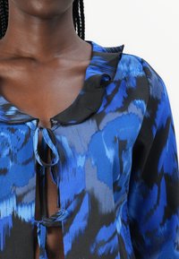 Blusa blu e nera a fantasia con scollatura arricciata e chiusura con fiocco, realizzata in un tessuto leggero e testurizzato. Vista ravvicinata.