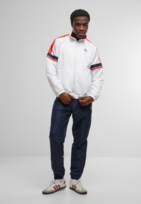 White zip-up jacket with red and navy stripes, paired with navy pants and white shoes featuring red accents. Smooth fabric, relaxed fit.