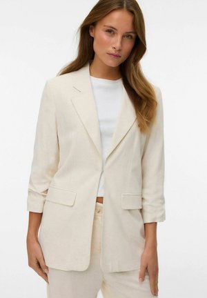 Femeie cu păr lung, purtând un sacou bej deschis peste o cămașă albă și pantaloni asortați, stând în fața unui fundal alb simplu.