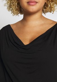 Nära bild av en person med lockigt blont hår som bär guldörhängen i form av hooper och en svart top med draperad hals mot en enfärgad bakgrund.