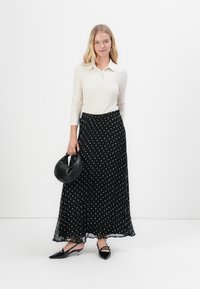 Gonna nera lunga plissettata con pois bianchi, top crema a maniche lunghe, scarpe con tacco nere e borsa a spalla nera.