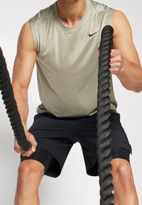 Khaki bezrękawnik sportowy o gładkiej fakturze, w połączeniu z czarnymi szortami z bocznymi akcentami. Użytkownik trzyma gruby, czarny linowy bojowy.