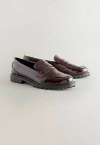 Mocassins en cuir verni marron avec une semelle en caoutchouc texturé, présentant une sangle décorative sur le devant et une finition lisse.