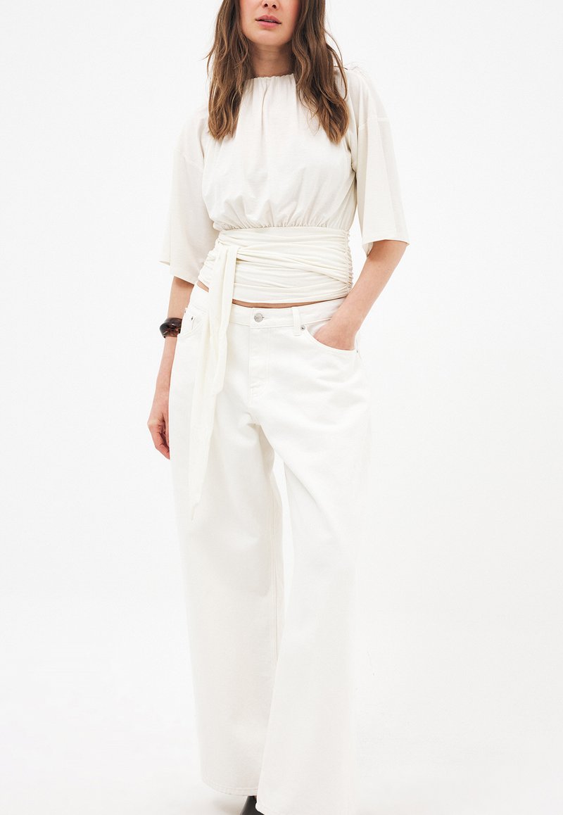 Mujer con blusa blanca holgada de estilo cruzado y pantalones blancos de pierna ancha, de pie con una mano en el bolsillo sobre un fondo liso.