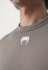 Barna pamut póló kerek nyakkal. A mellkason egy fehér pók grafika található. Simább textúra, rövid ujjú, minimalista dizájn.