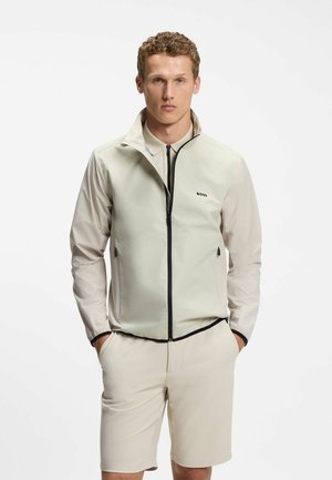Ung mand iført lys beige lynlåstrøje med "BOSS"-logo, khaki shorts, står med hænderne i lommerne mod ensfarvet baggrund.