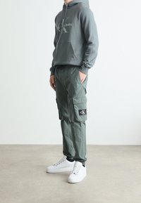 Szara bluza z kapturem z logo, zielone spodnie typu cargo z bocznymi kieszeniami i naszywką oraz białe trampki. Tkanina wydaje się miękka, design jest casualowy.