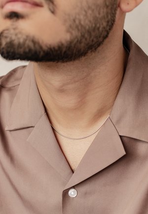 Silberne Halskette auf einem hellbraunen Shirt mit weicher Textur, das einen glänzenden Knopf hat. Nahaufnahme des Halsbereichs.
