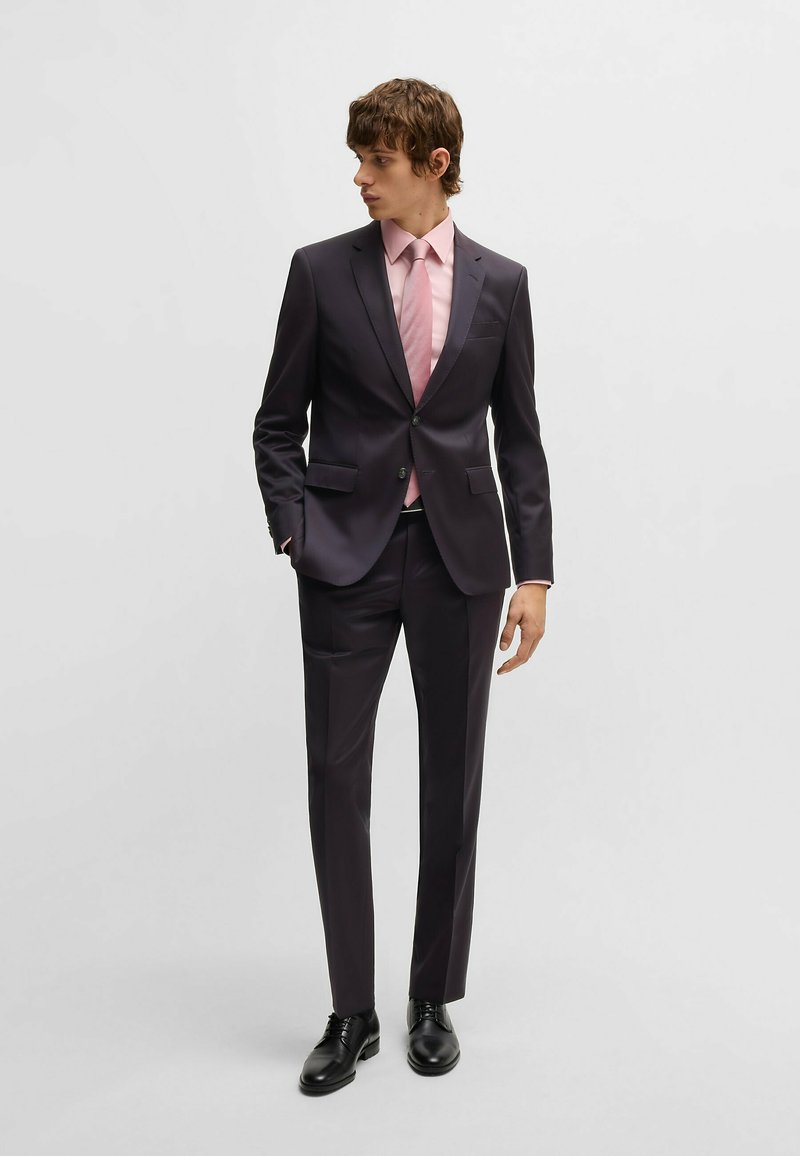 Donker op maat gemaakt kostuum met een getailleerd silhouet, gecombineerd met een lichtroze overhemd en stropdas. Glad materiaal, zakken op het jasje en de broek, zwarte schoenen.