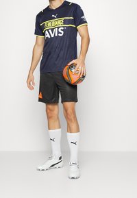 Merimustat, lyhythihaisat jalkapallopaidat, joissa on keltaisia yksityiskohtia, mustat shortsit, valkoiset sukat ja valkoiset kengät, pitelee oranssia ja mustaa jalkapalloa.