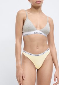 Bralette gris en forma de triángulo con tirantes finos, banda blanca con el logo de Calvin Klein; tanga amarillo a juego con una cinturilla blanca con logo. Textura suave.