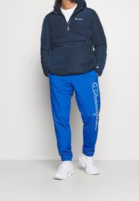 Chaqueta de pullover de color azul marino con media cremallera, bolsillo frontal, combinada con pantalones de chándal azul vibrante que presentan un estampado de logo blanco en el lateral. Zapatillas blancas.