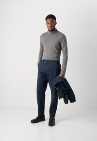 Sivi pulover s turtleneck-om, mornarsko plave krojene hlače i crne gležnjače. Model drži mornarsko plavi sakou. Glatke teksture, jednostavan dizajn.