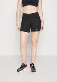 Svarta träningsshorts med hög midja, prydda med vit Puma-logga, slät textur och kort längd, kombinerade med svarta sneakers.