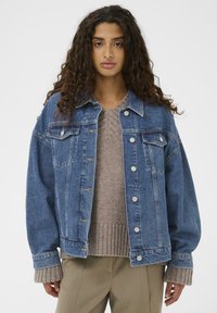 Traper jakna u tamnoplavoj boji sa srebrnim dugmadi na kopčanju, dva džepa na prsima i opuštenim krojem, složena preko bež pletene dukserice.