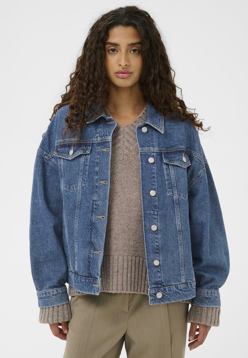 Traper jakna u tamnoplavoj boji sa srebrnim dugmadi na kopčanju, dva džepa na prsima i opuštenim krojem, složena preko bež pletene dukserice.