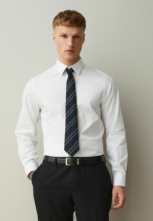 Young man wearing a white dress shirt, navy striped tie, black belt, and black trousers, standing against a plain light-colored wall.