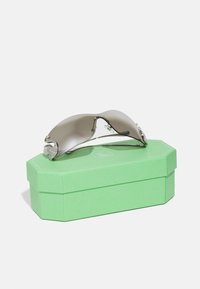 Sunglasses with a curved gray lens, silver accents, and crystal details on the temples, resting on a green octagonal box.