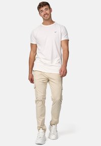 T-shirt blanche à manches courtes avec un petit logo, associée à un pantalon cargo beige avec des poches latérales, complétée par des baskets blanches.