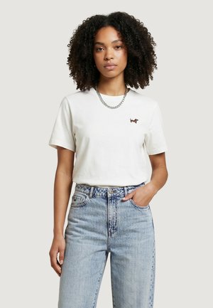 Mujer con el cabello rizado, vestida con una camiseta blanca con un pequeño logo de perro y unos vaqueros de tiro alto azul claro, de pie con una mano en el bolsillo.