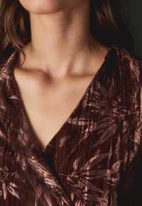 Braunes Samtoberteil mit V-Ausschnitt, das ein dezentes Blattmuster aufweist. Die Textur wirkt weich mit einem leichten Glanz.