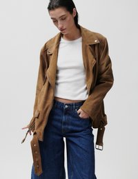 Γυναίκα που φοράει καφέ σουέντ μπουφάν πάνω από λευκό crop top και τζιν ψηλόμεσο, με το χέρι στην τσέπη, κοιτάζοντας προς τα κάτω.