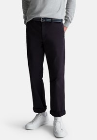 Hombre con pantalones oscuros navy con puños, zapatillas blancas, una camisa de manga larga gris y un cinturón trenzado azul con hebilla de metal.