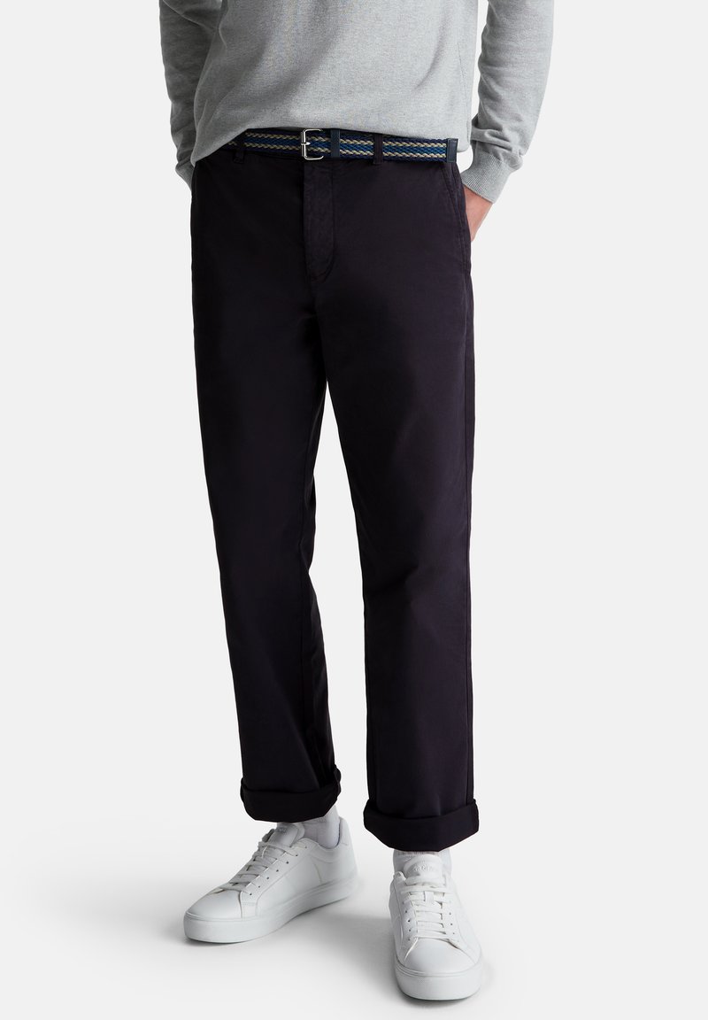 Hombre con pantalones oscuros navy con puños, zapatillas blancas, una camisa de manga larga gris y un cinturón trenzado azul con hebilla de metal.