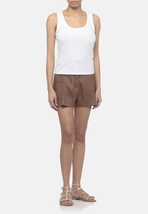 Donna che indossa un top bianco senza maniche, pantaloncini marroni a vita alta e sandali bassi beige con borchie, in piedi davanti a uno sfondo semplice.