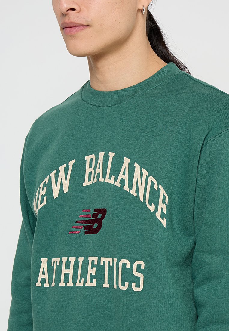 Felpa verde in cotone con colletto a girocollo. Scritte "NEW BALANCE" e "ATHLETICS" stampate in crema, con un accento di logo color bordeaux sotto.