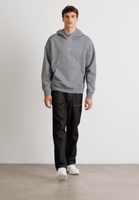 Grå hettegenser med en frontlomme, som har en liten logo. Kombinert med svarte jeans og hvite joggesko på et lyst gulv.