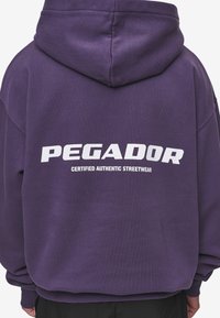 Felpa viola realizzata in tessuto morbido, con un grande logo bianco "PEGADOR" e la scritta "CERTIFIED AUTHENTIC STREETWEAR" sul retro.