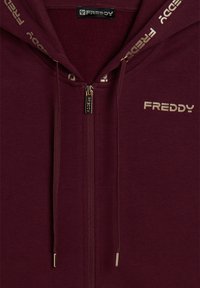 Felpa con zip color bordeaux, logo dorato, tessuto testurizzato e lacci a contrasto con punte metalliche. Cappuccio foderato con un materiale morbido.