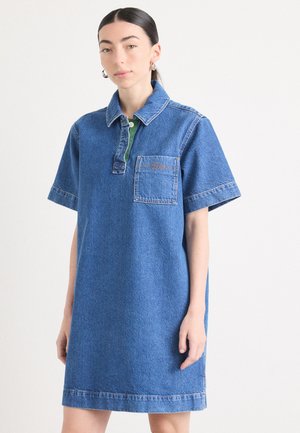 Robe en denim de couleur bleue avec un col, des manches courtes et une poche sur la poitrine. Elle présente des finitions vertes et des détails de couture contrastés.
